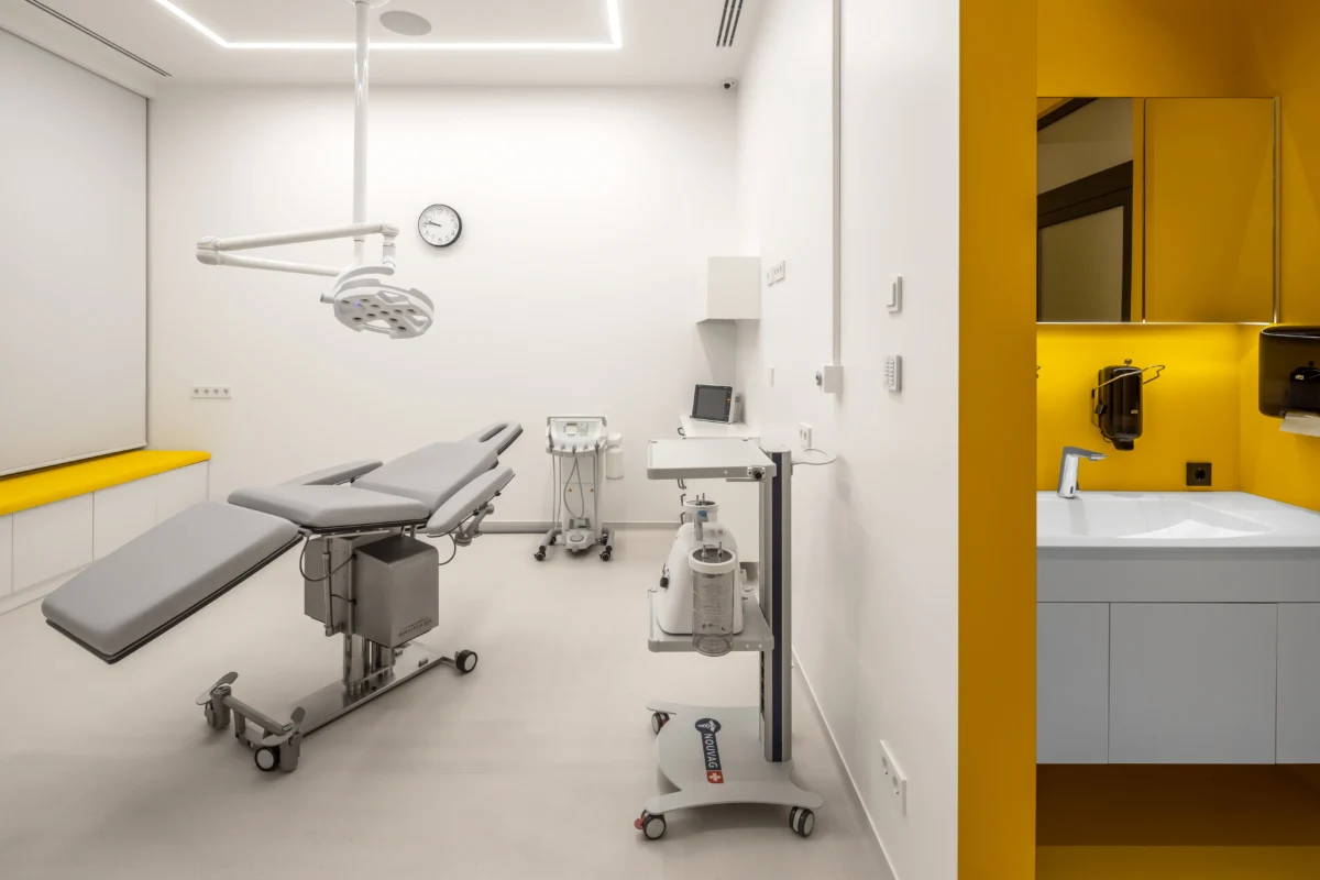 A modern medical examination room from SPACE DESIGN's operating room project features a gray examination chair in the center, an overhead light, and various medical equipment. The room has bright yellow accents, a wall clock, and a sink area with a mirror and soap dispenser. It maintains a clean and orderly environment reminiscent of an expertly designed SPACE DESIGN operating room project.