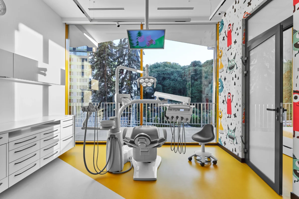 An inviting children's dental office by SPACE features their expertly crafted interior design, including a brightly lit space with a comfortable dental chair, advanced dental equipment, and a mounted monitor showcasing a cartoon. The white walls are adorned with colorful monster decals, while large windows offer views of trees and buildings outside.