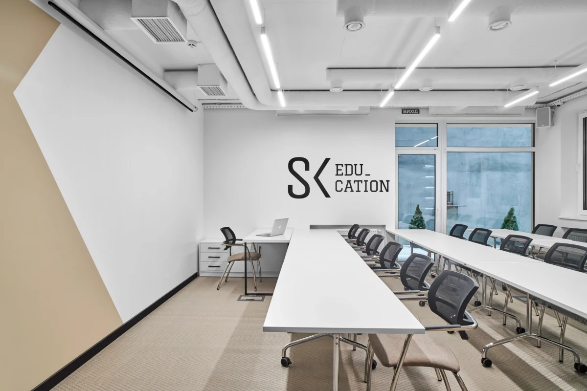 A modern, white minimalist classroom with long tables and multiple chairs facing a whiteboard and a wall. A laptop sits on one of the tables near the wall with the word  Solonko Sovyak "Education" creatively written on it. Bright ceiling lights illuminate the space.