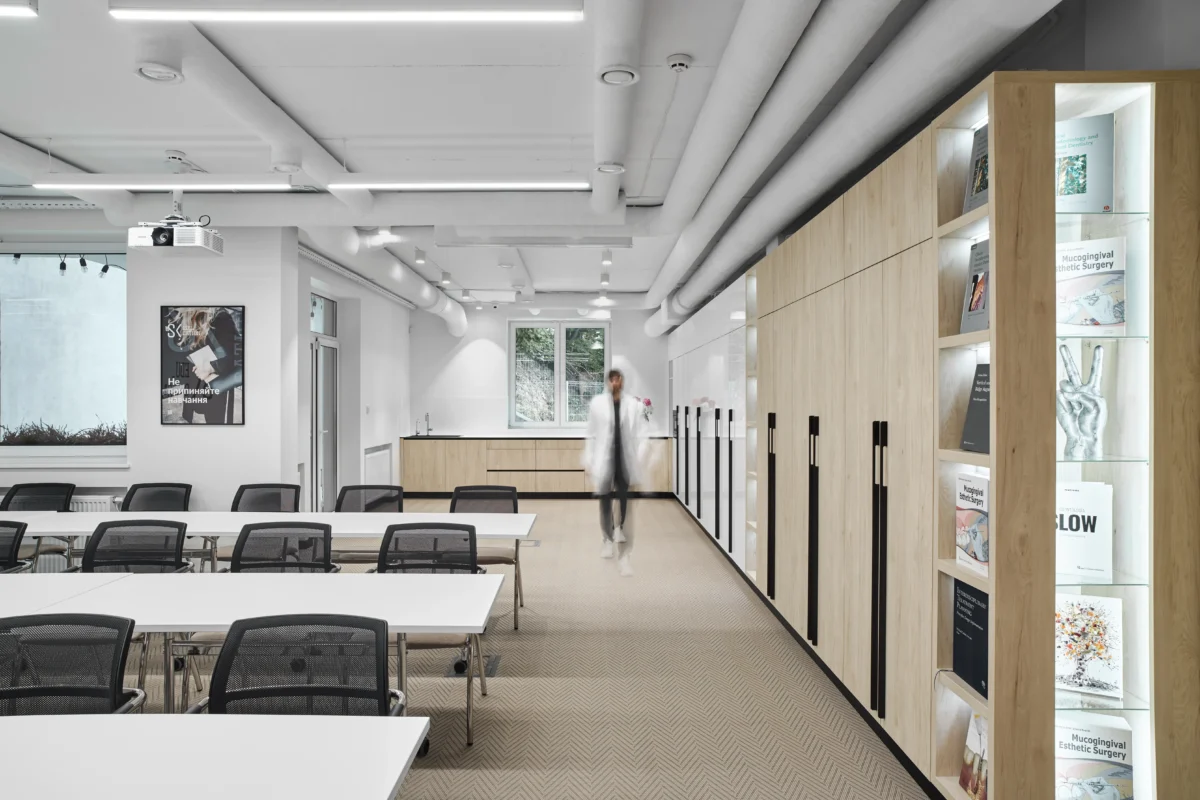 A modern conference room with white tables and black chairs lined up facing a projection screen. A person in white clothing walks past a wall-mounted cabinet filled with magazines and books. Large windows and framed posters decorate the well-lit space.