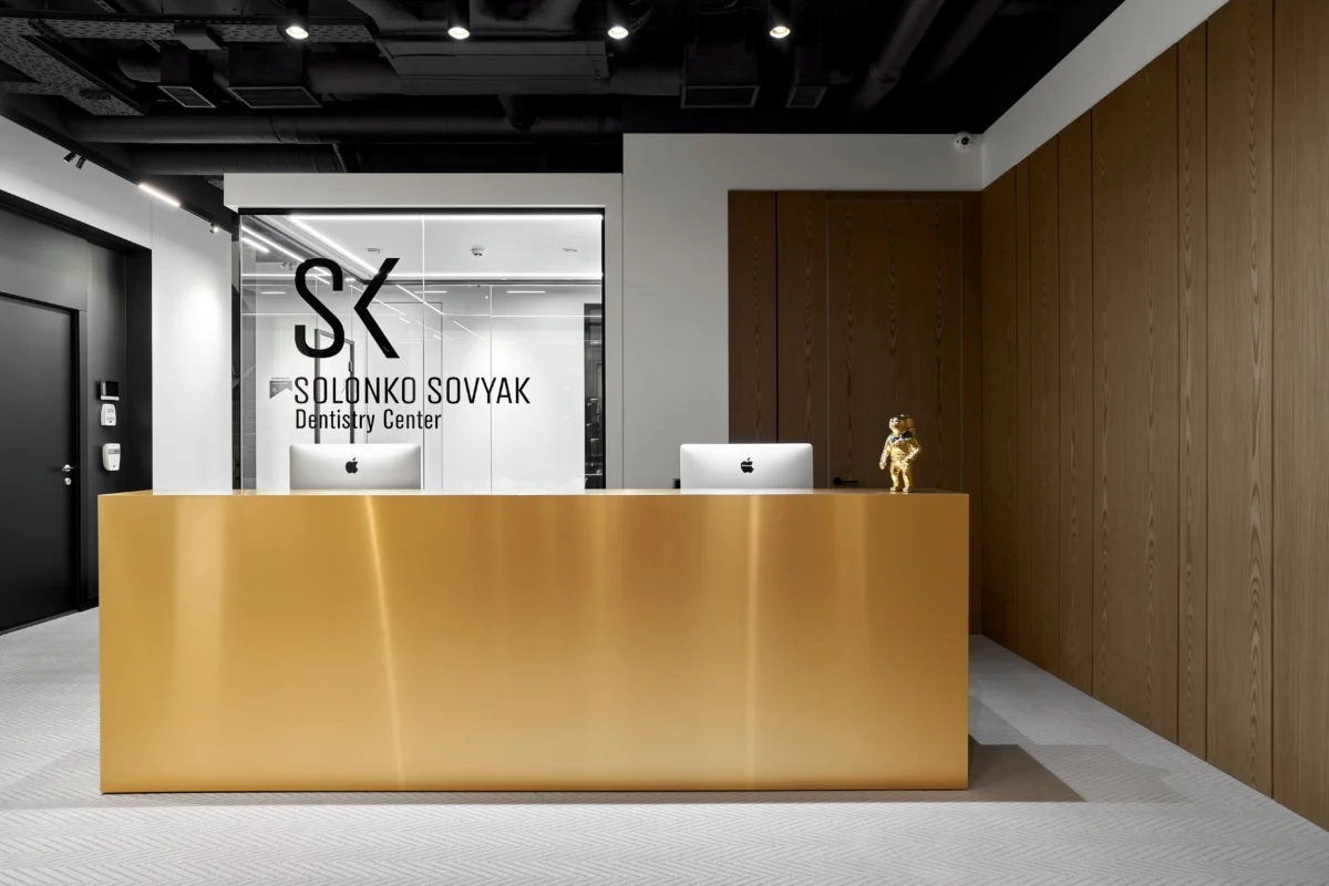 Modern dentistry center front desk area with a sleek golden reception counter. Two computer monitors are on the counter, with branding "Solonko Sovyak Dentistry Center" visible behind. The space features minimalistic decor with wooden panels and monochromatic color scheme.