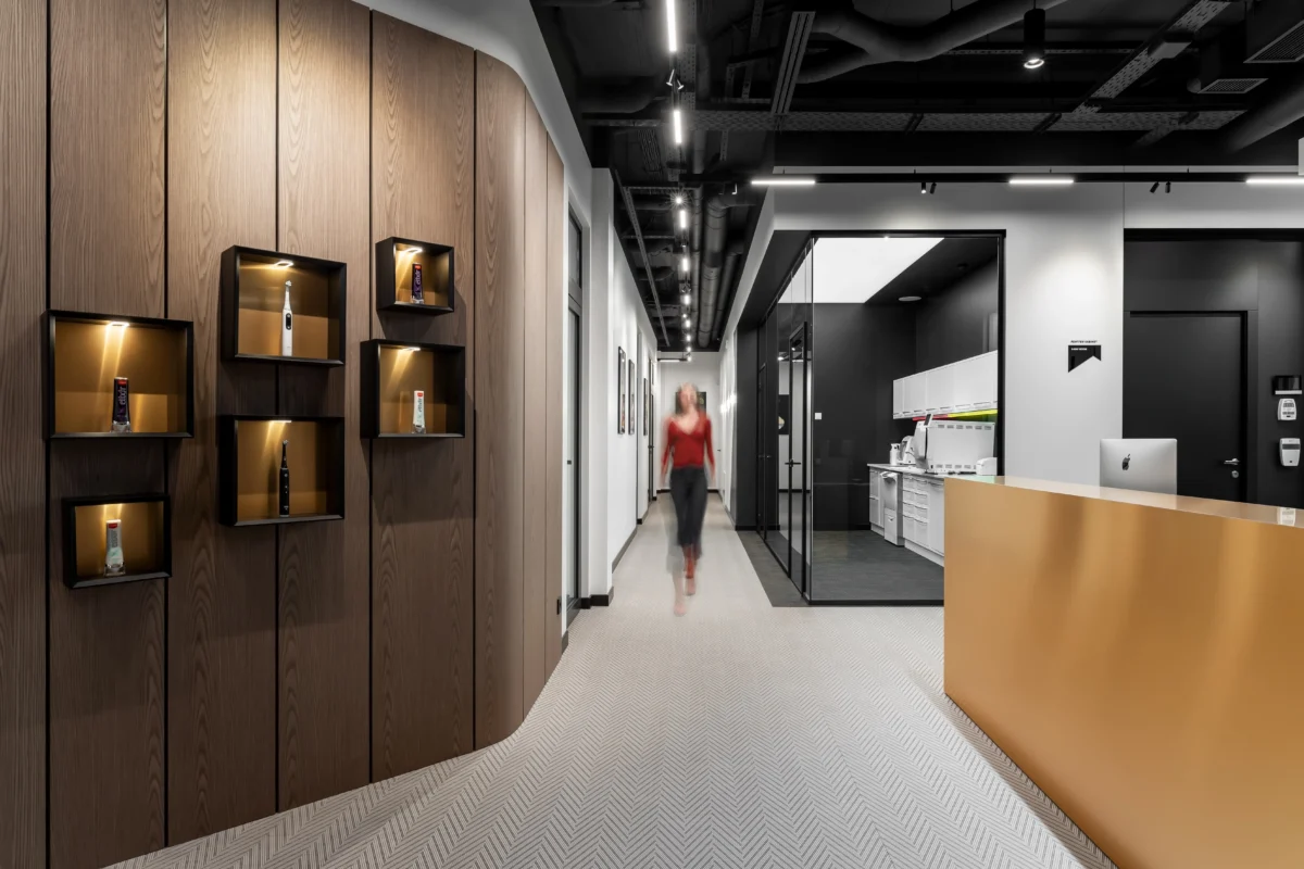 A modern dental center corridor optimized for a dental clinic, featuring SPACE DESIGN's "dentistry project" with a wooden display wall incorporating illuminated shelves holding toothbrush items. A blurred figure walks down the middle of the corridor. To the right, there is a sleek reception desk in front of a glass-walled room that appears to be sterilization room.