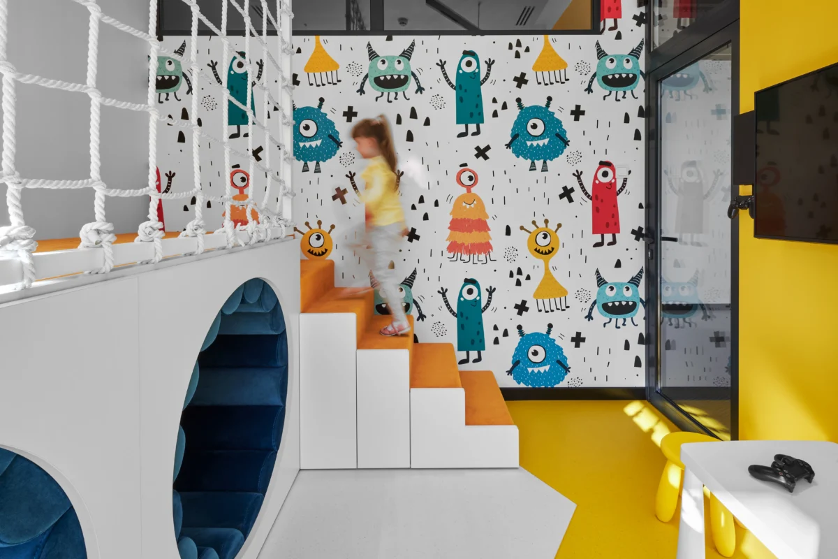A child is ascending orange stairs in a colorful playroom adorned with a playful monster-themed wall mural, characteristic of dental offices for children. The space, designed by SPACE DESIGN's interior design team, includes a rope climbing area, a navy blue cushioned nook, and a TV mounted on a yellow wall. Vibrant colors and whimsical designs enhance the fun atmosphere.