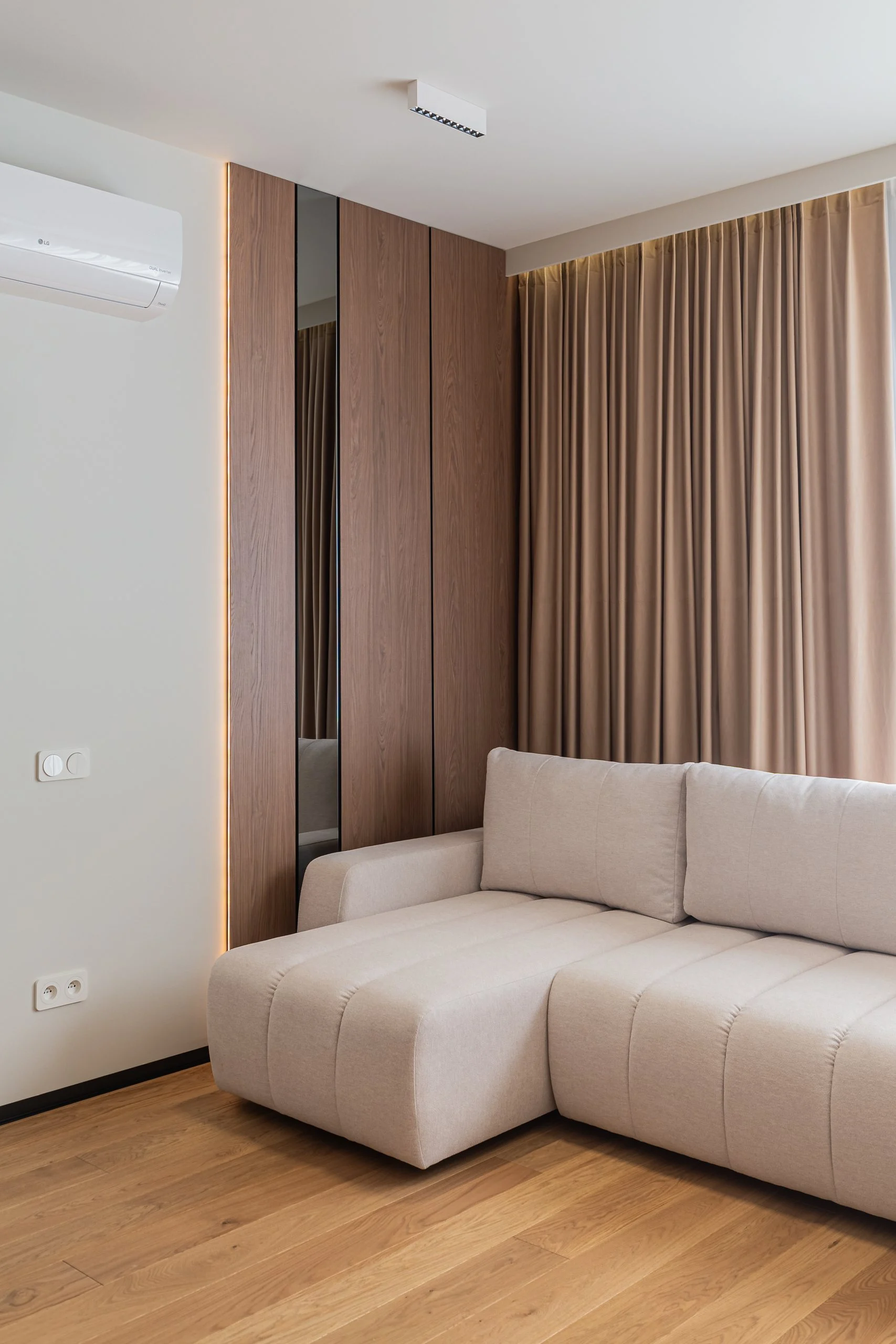 Beżowa sofa narożna przy oknie z beżowymi zasłonami i drewnianym panelem