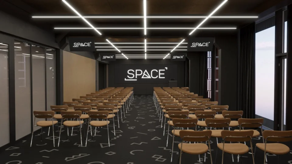 Conference hall with theatre-style seating: two blocks of chairs, a lit logo media wall and linear ceiling lights